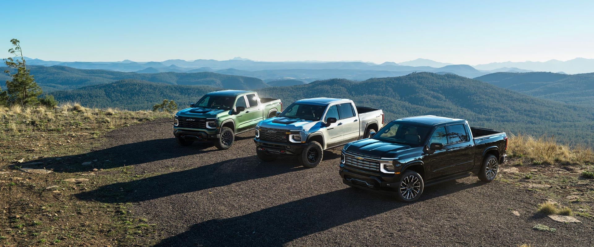 Three 2027 Chevrolet Silverado 1500 pickup trucks (green, black) parked in a row on a dirt field with a mountain range backdrop.